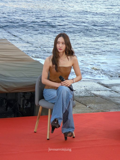 Jung Soojung sitting on a chair holding a microphone. She wears a strapless brown top, blue jeans, and black heels. The setting includes a red carpet, ocean waves, and a concrete pier.