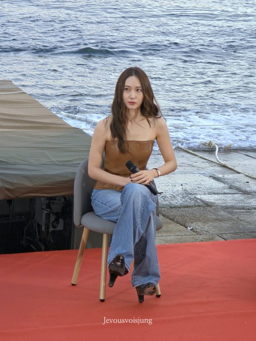 Jung Soojung sitting on a chair holding a microphone. She wears a strapless brown top, blue jeans, and black heels. The setting includes a red carpet, ocean waves, and a concrete pier.