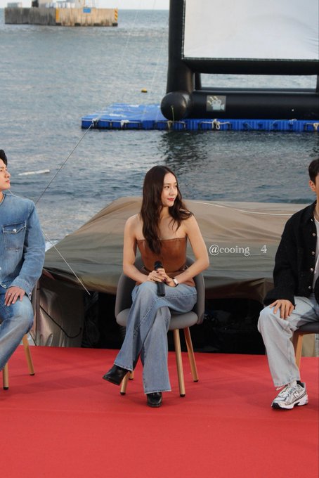 Jung Soojung standing on a red carpet holding a microphone, wearing a brown strapless top and wide-leg blue jeans, with long brown hair. She is positioned in front of a body of water and a large screen, with two other individuals seated nearby. A watermark reading "@cooing__4" is visible.