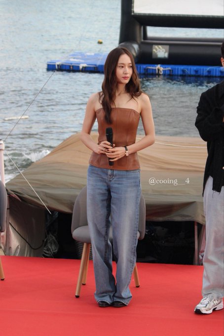 Jung Soojung standing on a red carpet holding a microphone, wearing a brown strapless top and wide-leg blue jeans, with long brown hair. She is positioned in front of a body of water and a large screen, with two other individuals seated nearby. A watermark reading "@cooing__4" is visible.