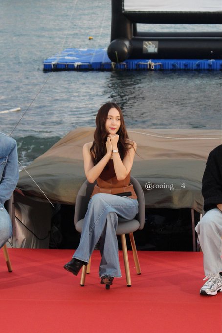 Jung Soojung standing on a red carpet holding a microphone, wearing a brown strapless top and wide-leg blue jeans, with long brown hair. She is positioned in front of a body of water and a large screen, with two other individuals seated nearby. A watermark reading "@cooing__4" is visible.