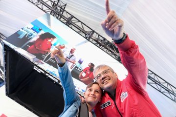 A man in a red jacket with a name tag pointing upward, standing next to a person in a blue jacket, with a large screen displaying people in red jackets behind them. A crowd gathers around a display of two large rocket models. A group of people in red and black jackets stands on a stage with a sign reading "TEKNOFEST BLOKZİNCİR YARIŞMASI." People browse booths under a tent with banners displaying "TÜBİTAK" and "TEKNOFEST."
