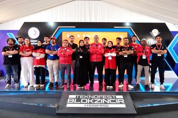 A man in a red jacket with a name tag pointing upward, standing next to a person in a blue jacket, with a large screen displaying people in red jackets behind them. A crowd gathers around a display of two large rocket models. A group of people in red and black jackets stands on a stage with a sign reading "TEKNOFEST BLOKZİNCİR YARIŞMASI." People browse booths under a tent with banners displaying "TÜBİTAK" and "TEKNOFEST."