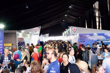 A man in a red jacket with a name tag pointing upward, standing next to a person in a blue jacket, with a large screen displaying people in red jackets behind them. A crowd gathers around a display of two large rocket models. A group of people in red and black jackets stands on a stage with a sign reading "TEKNOFEST BLOKZİNCİR YARIŞMASI." People browse booths under a tent with banners displaying "TÜBİTAK" and "TEKNOFEST."