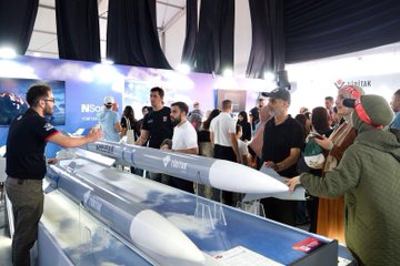 A man in a red jacket with a name tag pointing upward, standing next to a person in a blue jacket, with a large screen displaying people in red jackets behind them. A crowd gathers around a display of two large rocket models. A group of people in red and black jackets stands on a stage with a sign reading "TEKNOFEST BLOKZİNCİR YARIŞMASI." People browse booths under a tent with banners displaying "TÜBİTAK" and "TEKNOFEST."