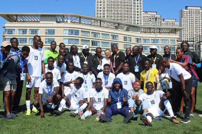 A group of football players in white jerseys with numbers, some wearing green, posing together on a grassy field. Several individuals wear lanyards with badges. Tall buildings and a clear blue sky are visible in the background.