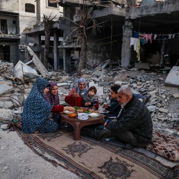 A long table set for a formal dinner in an ornate room with red carpet, wooden paneling, and chandeliers, filled with people in formal attire. The table is decorated with flowers, candles, and place settings. A family sits on a patterned rug eating from a small table amidst rubble and damaged buildings, with palm trees and laundry visible in the background.
