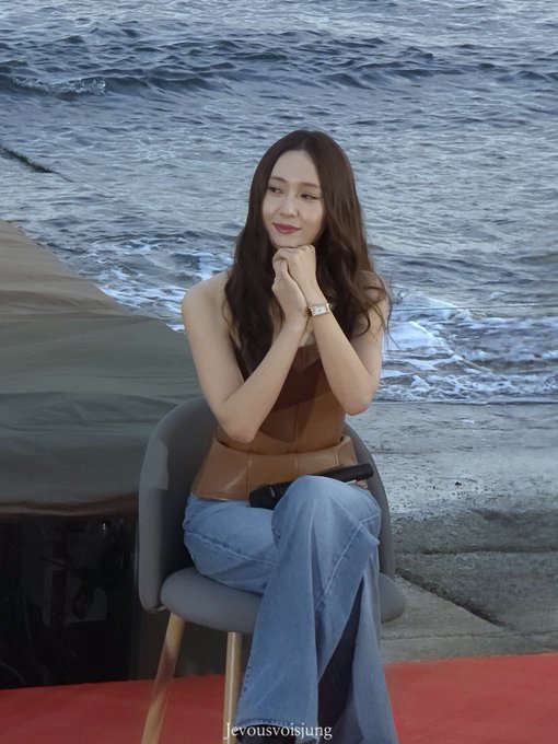 Jung Soojung sitting on a chair by the ocean, wearing a brown strapless top and blue flared jeans, holding a microphone, with long dark hair flowing over her shoulders.