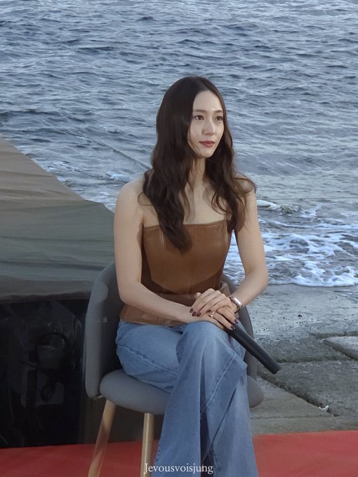 Jung Soojung sitting on a chair by the ocean, wearing a brown strapless top and blue flared jeans, holding a microphone, with long dark hair flowing over her shoulders.