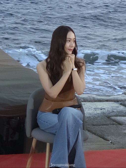 Jung Soojung sitting on a chair by the ocean, wearing a brown strapless top and blue flared jeans, holding a microphone, with long dark hair flowing over her shoulders.