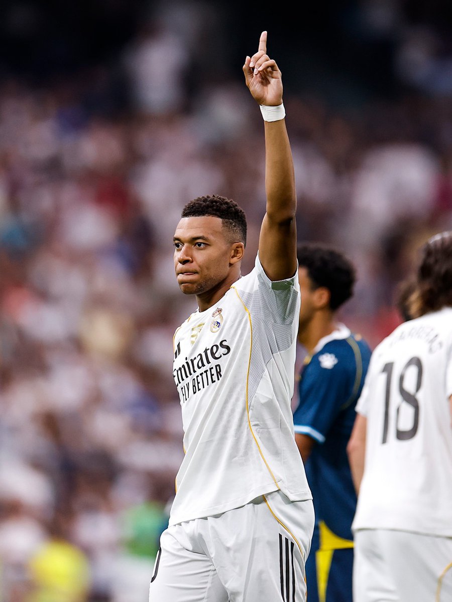 Jude Bellingham wearing a white Real Madrid jersey with the Emirates "Fly Better" sponsor logo, raising his right arm with one finger pointing upward. He is on a soccer field with other players in the background, one wearing a dark blue jersey with the number 18.