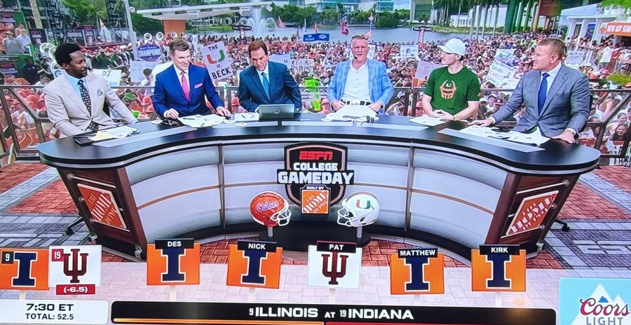 A panel of commentators on a curved desk with ESPN College GameDay branding. Visible individuals include Pat McAfee and others in suits or casual attire. The desk displays logos for Illinois and Indiana, with a football graphic. Text banners show "ILLINOIS at INDIANA" and team scores.