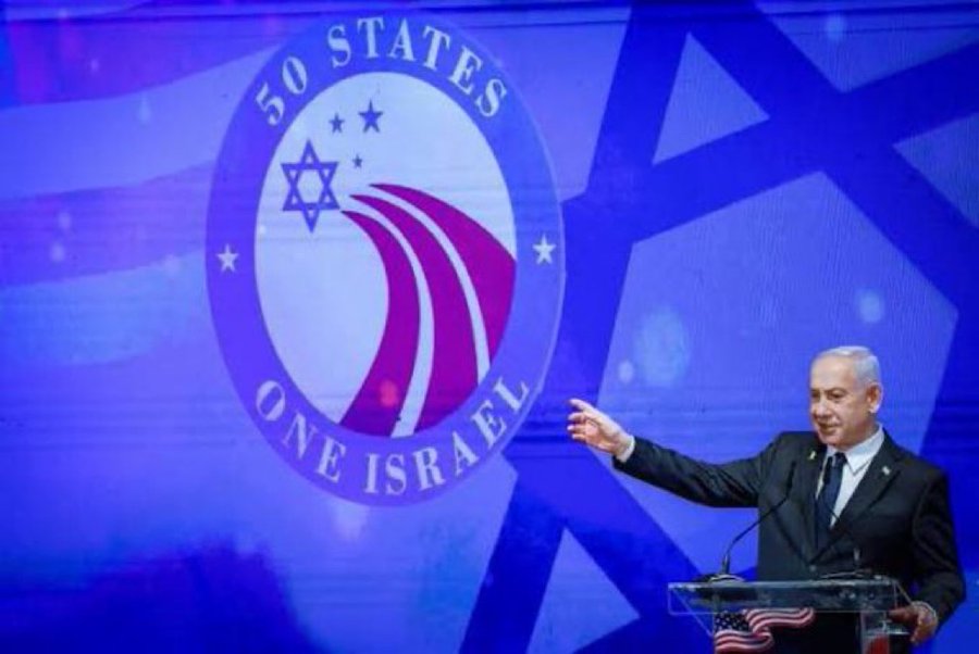 Benjamin Netanyahu standing at a podium with an American flag, gesturing with his right hand. Behind him, a large screen displays a logo with "50 STATES ONE ISRAEL," featuring a Star of David, purple and white stripes, and stars.