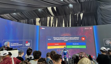 Two children in red jackets with Türkiye Uzay Ajansı logos hold rocket models. People in a large tent with blue drapes and tables engage in activities. A screen displays a question about astronauts. Individuals use virtual reality headsets and interact with technology displays.