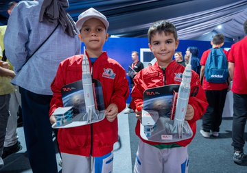 Two children in red jackets with Türkiye Uzay Ajansı logos hold rocket models. People in a large tent with blue drapes and tables engage in activities. A screen displays a question about astronauts. Individuals use virtual reality headsets and interact with technology displays.