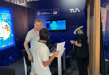 Two children in red jackets with Türkiye Uzay Ajansı logos hold rocket models. People in a large tent with blue drapes and tables engage in activities. A screen displays a question about astronauts. Individuals use virtual reality headsets and interact with technology displays.