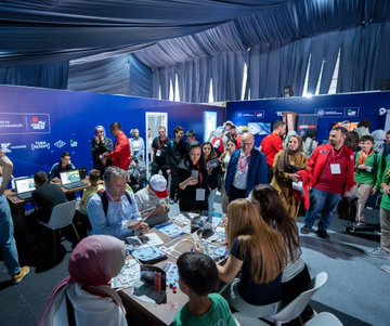 Two children in red jackets with Türkiye Uzay Ajansı logos hold rocket models. People in a large tent with blue drapes and tables engage in activities. A screen displays a question about astronauts. Individuals use virtual reality headsets and interact with technology displays.