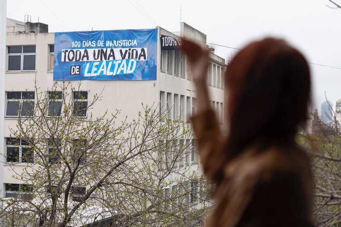 A person with reddish-brown hair wearing a hooded jacket, raising one hand, stands in the foreground. In the background, a building displays a large blue and white banner with text reading "100 DÍAS DE INJUSTICIA TODA UNA VIDA DE LEALTAD".