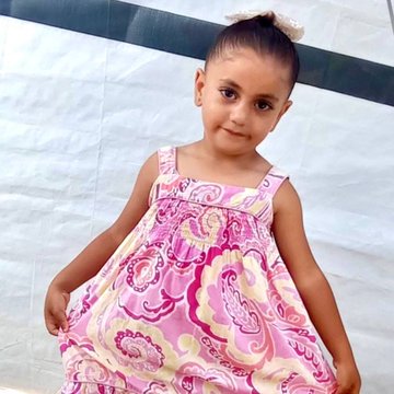 Mohammed Zuhair, a boy wearing a yellow cap and green shirt with pockets, smiling and raising a hand. Khadija, a girl in a pink and yellow patterned dress with a bow in her hair, standing and holding her dress. Both are in an outdoor setting with a dirt ground and a tent visible in the background.