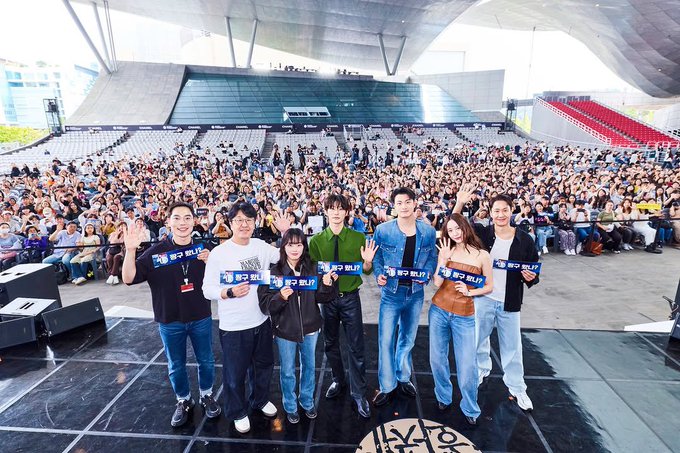 A woman with long brown hair wearing a strapless brown top and blue jeans sits at a white table holding a microphone, smiling. A water bottle and mug are on the table. Behind her, a digital screen displays partial text including "BIFF" and "Audition109." A group of people stands on a stage in a large outdoor stadium with a crowd, holding blue banners with text, wearing casual clothing like jeans and jackets.