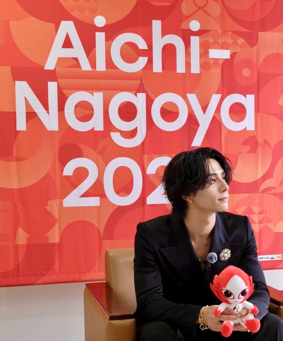 Nishi Hiroto sitting on a chair, wearing a black suit and holding a red and white plush toy. A backdrop behind him displays the text "Aichi-Nagoya 2026" in large white letters on a red background with circular patterns.