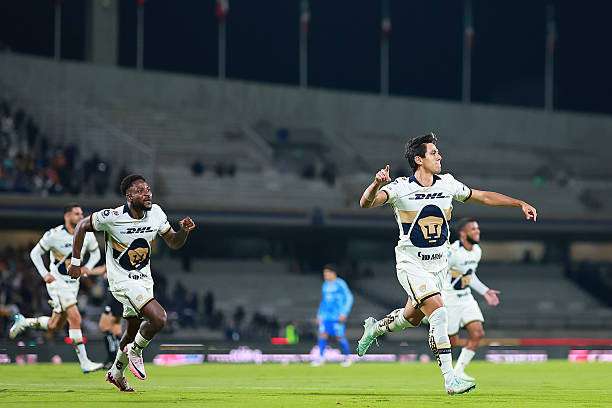 Soccer players in Pumas jerseys with DHL and Puma logos running on a grassy field in a stadium. José Juan Macías and other players are visible, celebrating with raised arms. The stadium has tiered seating with some spectators and flags in the background.