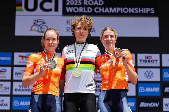 Marlen Reusser wearing a white jersey with rainbow stripes and a gold medal around her neck, standing on a podium. Anna van der Breggen and Demi Vollering, both in orange jerseys, stand beside her, each wearing silver and bronze medals. A backdrop displays "UCI 2025 ROAD WORLD CHAMPIONSHIPS KIGALI RWANDA" and various sponsor logos.