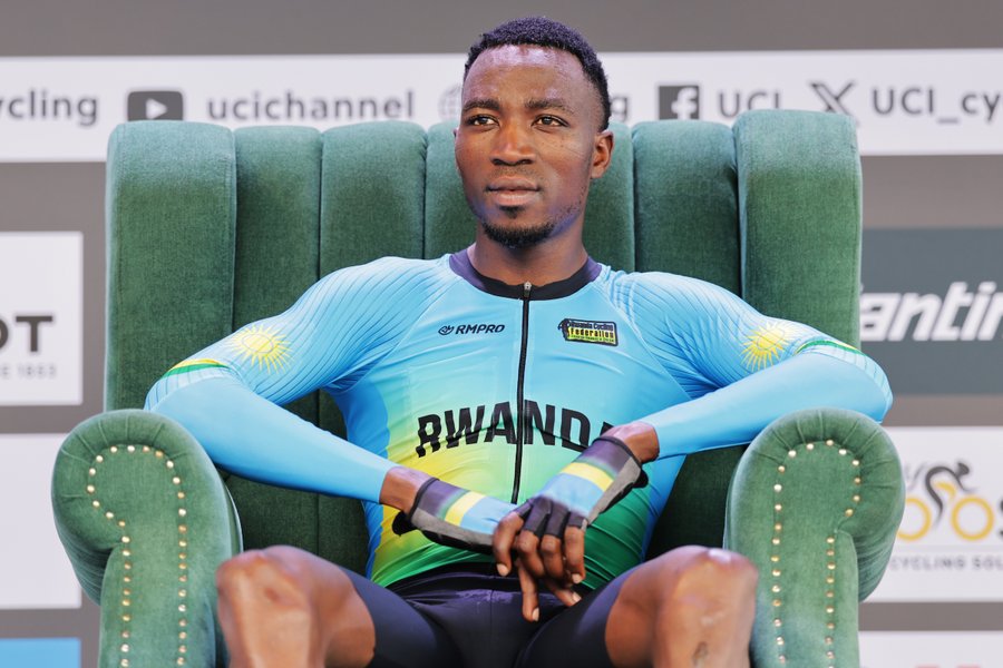 Shemu Nsengiyumva sitting in a green upholstered chair, wearing a blue cycling jersey with "RWANDA" and yellow accents, black cycling shorts, and gloves. The background includes banners with "UCI" and "Kigali2025" text.