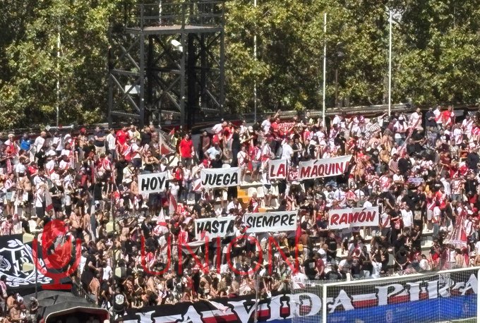 A crowd of fans at a soccer stadium, many wearing white and red clothing. Several large banners display text, including 
