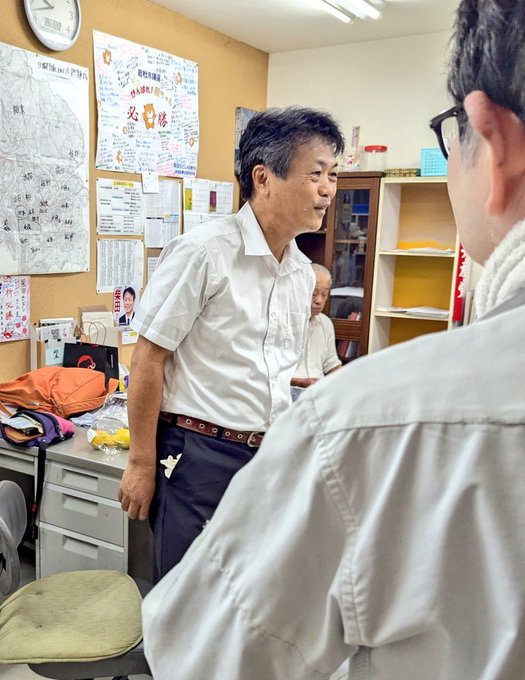 Satoshi Shibata wearing a white short-sleeve shirt and dark pants, standing in an office with beige walls. Posters and papers are pinned to the walls, and shelves with books and files are visible. Another person in a white shirt is partially visible, facing Satoshi Shibata.