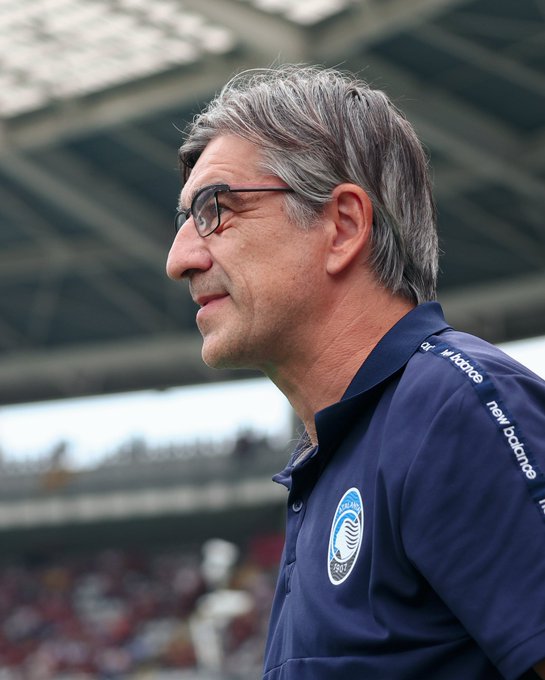 A man with gray hair and glasses, wearing a navy blue polo shirt with a circular logo and "New Balance" text on the sleeve, stands in a stadium with a crowd and a covered stand in the background.