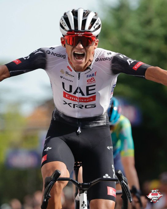Isaac del Toro montando en bicicleta, vistiendo una camiseta de ciclismo blanca y negra con los logotipos "UAE" y "XRG", pantalones cortos negros y un casco blanco con rayas negras. Gafas de sol rojas cubren sus ojos y sus brazos están extendidos. Una pequeña marca de agua de Instagram es visible en la esquina inferior derecha.