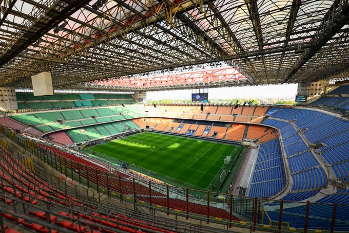 A large soccer stadium with green field, red, green, blue, and orange seating, and a metal roof structure. The stands are mostly empty, and the field is well-maintained.