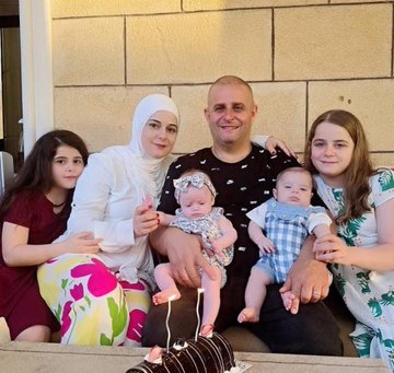 Two photos of a family. The first shows Shadi Sobhi Sharara, a man with a shaved head, sitting with Celine Shadi Sharara, a woman in a white hijab and floral pants, holding a baby in a floral outfit, and two girls, one in a red dress and one in a floral dress, with a baby in a blue outfit on Shadi\'s lap, next to a chocolate cake. The second shows four children, including a girl in a blue dress held by an older girl in a floral dress, a boy in a white shirt with a cartoon, and another boy, with heart emojis above their heads.