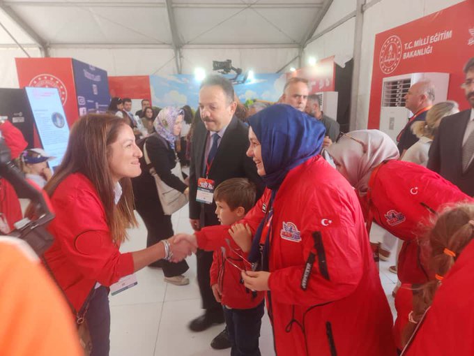 Nazif Yılmaz and Celile Eren Ökten standing at a booth with red panels displaying the Millî Eğitim Akademisi Başkanlığı logo. The booth features books, brochures, and a display of circular objects. People in red jackets and a child are visible, interacting with the booth. A backdrop shows an aerial image and text in Turkish.