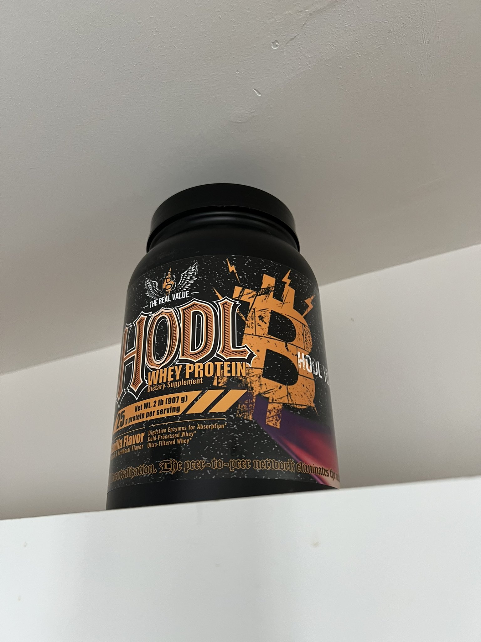 A photo of a HODL THE WHEY protein tub standing on a white kitchen shelf.