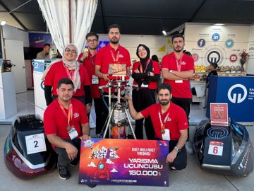 A group of people wearing red shirts and lanyards, standing and kneeling on a stage with a rocket model. They hold a trophy and a sign reading "Teknofest Dikey İnişli Roket Yarışması 3. Yarısmaya Katılım 150.000." Banners with "Makine ve Kimya Endüstrisi 75 Years" and "Transpera" are visible. Two numbered racing helmets are on the floor.