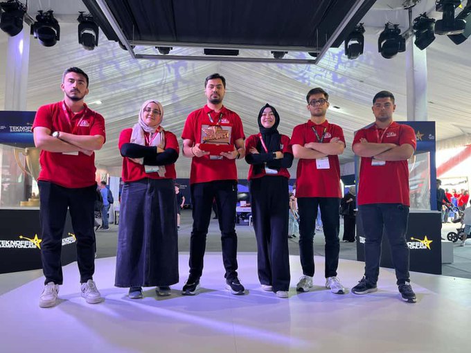 A group of people wearing red shirts and lanyards, standing and kneeling on a stage with a rocket model. They hold a trophy and a sign reading "Teknofest Dikey İnişli Roket Yarışması 3. Yarısmaya Katılım 150.000." Banners with "Makine ve Kimya Endüstrisi 75 Years" and "Transpera" are visible. Two numbered racing helmets are on the floor.