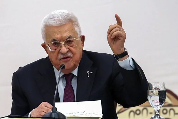 Mahmoud Abbas, wearing a dark suit, white shirt, and red tie, sits at a table with a microphone, gesturing with his right hand raised. Papers and a glass of water are on the table. Arabic text is visible on the papers and a banner in the background.