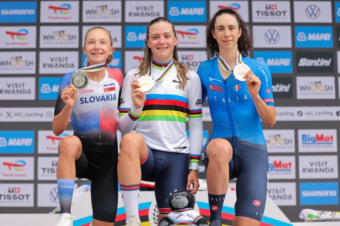 Zoe Backstedt, Viktoria Chladoñova, and Federica Venturelli on a podium, each wearing cycling uniforms and holding medals. Zoe Backstedt wears a rainbow-striped jersey, Viktoria Chladoñova a red and blue outfit with "Slovakia" text, and Federica Venturelli a blue outfit with "Italy" text. They are seated, with a stuffed animal on the podium.