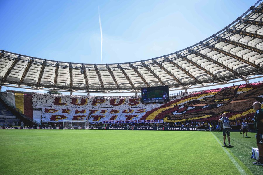 A large stadium with a green soccer field. Fans in the stands display a banner with "LUPUS PETITUS" in yellow and red letters, surrounded by colorful designs. A scoreboard and roof structure are visible under a clear blue sky. People are on the field near the sidelines.