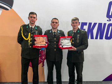 Three individuals in military uniforms holding award boxes, standing on a stage. A group of individuals in military uniforms posing together on a stage with a blue and white backdrop. Two people in casual attire holding a large check, flanked by individuals in military uniforms on a stage. Two individuals in business attire and military uniforms standing near a table with a sign labeled "INSPECTOR" and a screen displaying text.