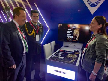 Three individuals in military uniforms holding award boxes, standing on a stage. A group of individuals in military uniforms posing together on a stage with a blue and white backdrop. Two people in casual attire holding a large check, flanked by individuals in military uniforms on a stage. Two individuals in business attire and military uniforms standing near a table with a sign labeled "INSPECTOR" and a screen displaying text.