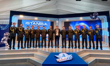 Three individuals in military uniforms holding award boxes, standing on a stage. A group of individuals in military uniforms posing together on a stage with a blue and white backdrop. Two people in casual attire holding a large check, flanked by individuals in military uniforms on a stage. Two individuals in business attire and military uniforms standing near a table with a sign labeled "INSPECTOR" and a screen displaying text.