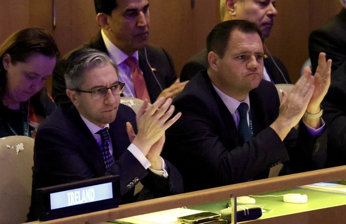 Simon Harris and another man in suits clapping at a formal meeting. A nameplate reading "Ireland" is visible on the table in front of them.