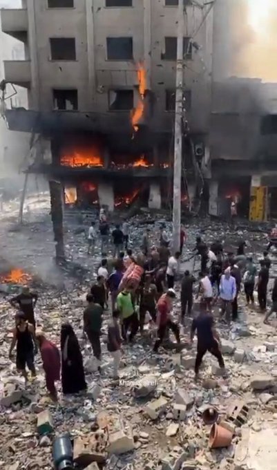 A multi-story building on fire with flames and smoke rising from the structure. Debris and rubble cover the ground, with people running and carrying injured individuals on stretchers. A firefighter in an orange vest and boots sprays water on the burning building. The scene shows chaos and destruction with a crowd gathered amidst the wreckage.