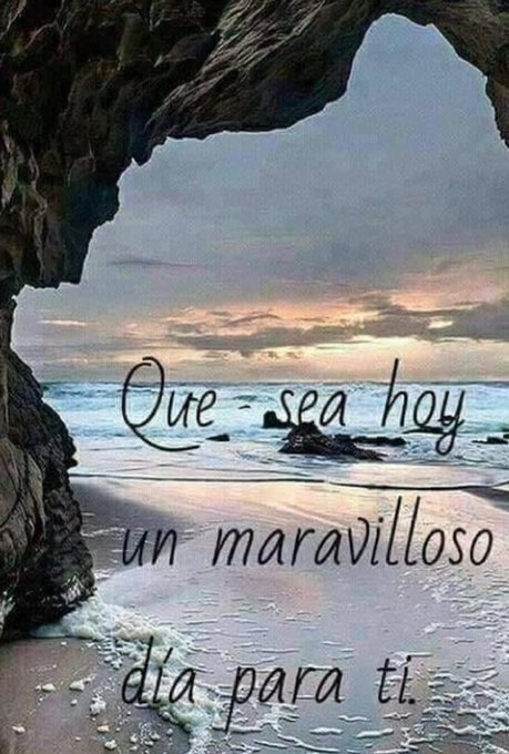 A beach scene with waves gently crashing on the shore. Rocky cliffs frame the left side, and the ocean extends to the horizon. A sunset sky with clouds casts a warm glow over the water and sand. Text overlay reads "Que sea hoy un maravilloso día para ti."