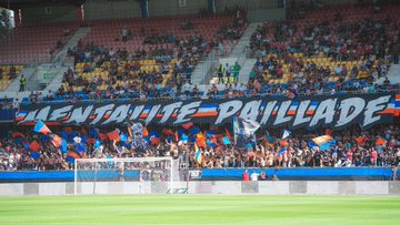 A crowd of fans in a stadium waving flags and banners. The banners display "RIBUNE POPULAIRE" in large orange and white letters and "MENTALITE PAILLADE" in black letters. A black flag with a white skull and "MONTPELLIER" is visible. The scene includes a soccer goal and field in the foreground, with fans raising their arms and cheering.