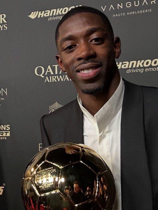 A man in a dark suit and white shirt holding a golden soccer ball trophy. The background features logos of sponsors including Qatar Airways and Hankook.