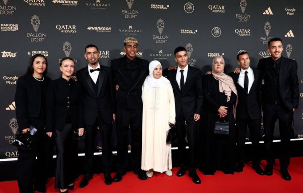Lamine Yamal stands in the center of a group on a red carpet, wearing a black suit. Surrounding him are family members, including women in elegant dresses and headscarves, and men in formal black suits and ties. The backdrop features logos of Qatar Airways, Ballon d\'Or, and other sponsors.